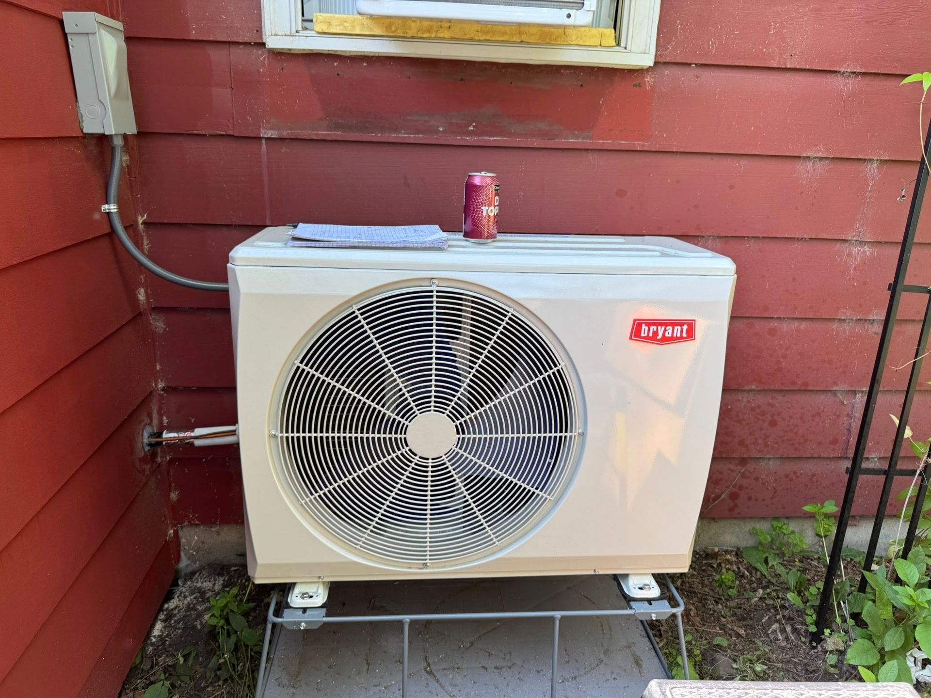 Outdoor Bryant air conditioner unit, beige, mounted on a metal stand, against a red wall with a window.
