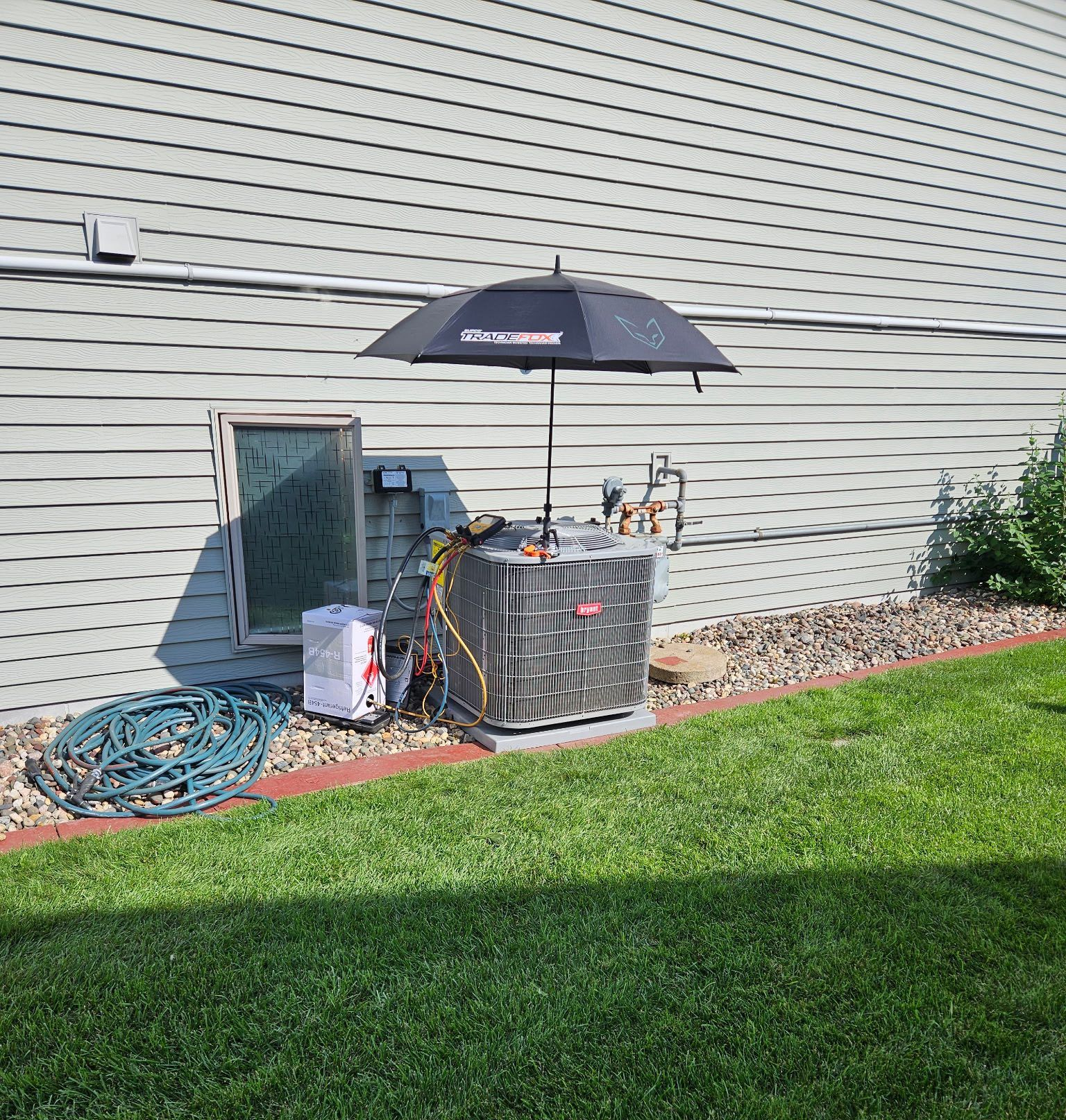 HVAC unit on a lawn, under an umbrella. Hoses and equipment surround the unit, next to a building with light siding.