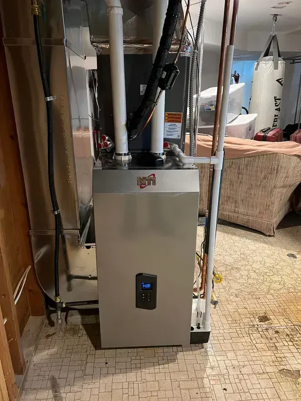 A silver furnace with a digital display installed in a basement. Copper pipes and ductwork are visible.