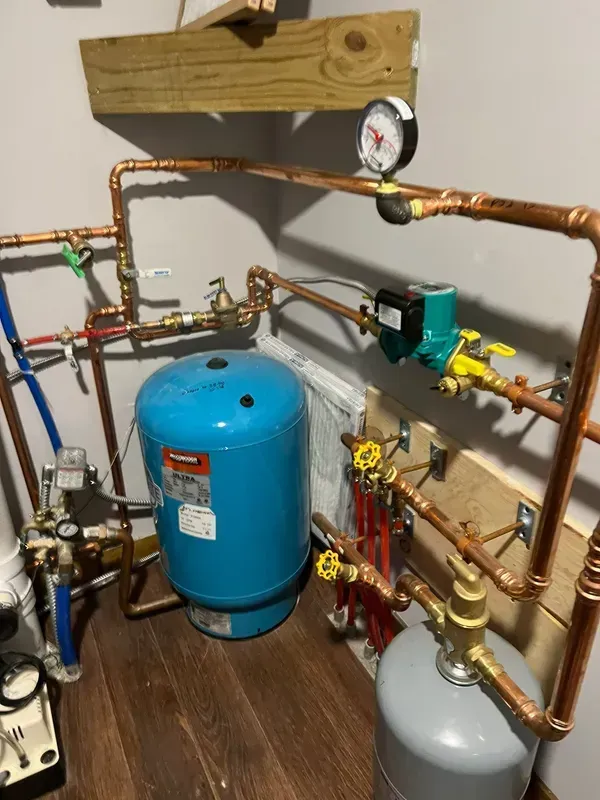 Copper pipes and tanks of a boiler system in a corner. Pressure gauge visible, wood beams overhead.
