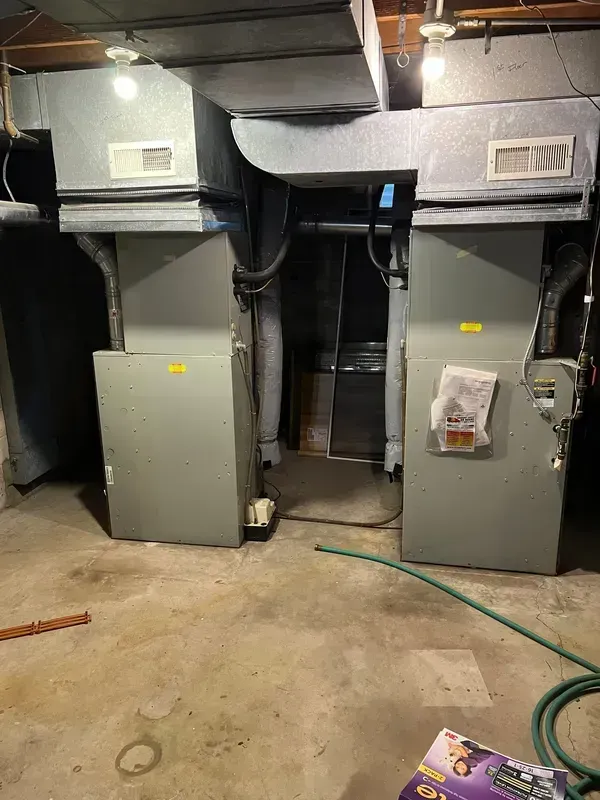 Two gray HVAC units side-by-side in a concrete basement. Ductwork and exposed pipes are visible.