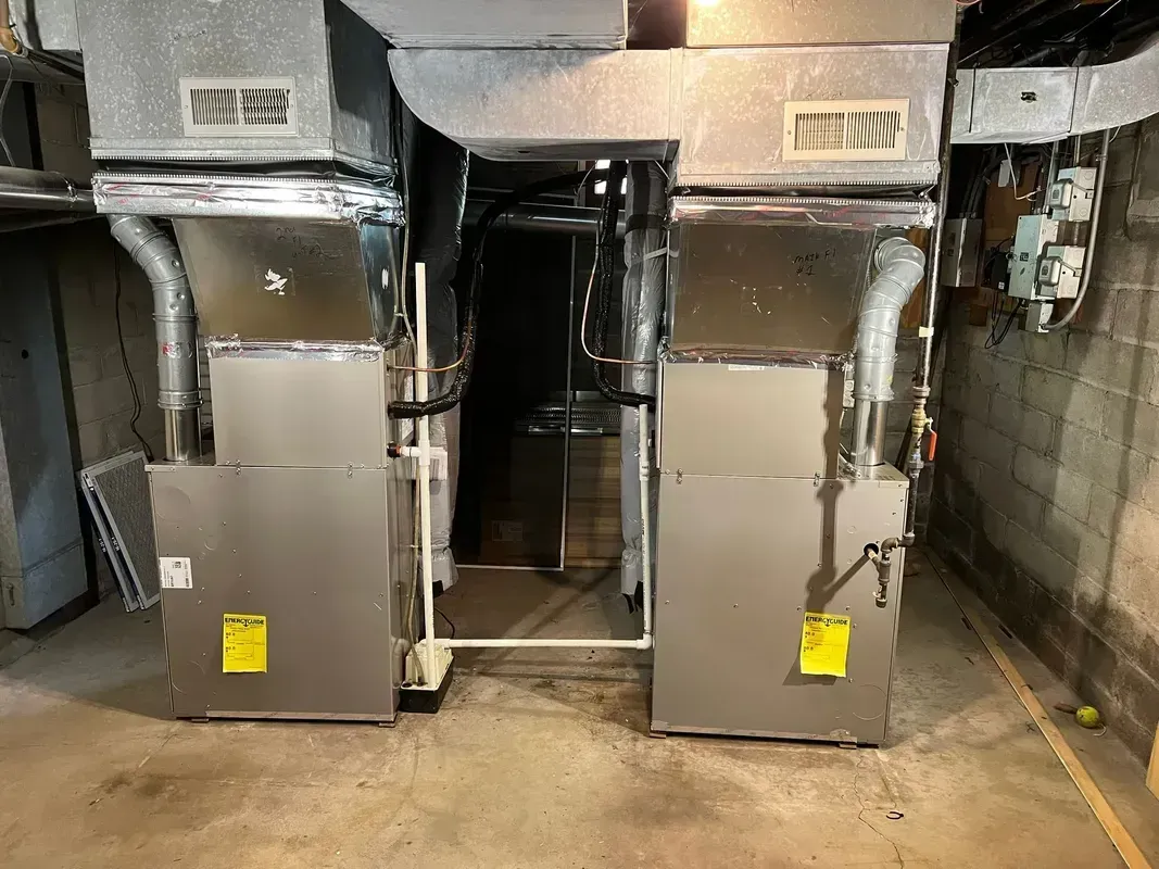 Two silver HVAC units in a basement with ductwork, set against a concrete wall.
