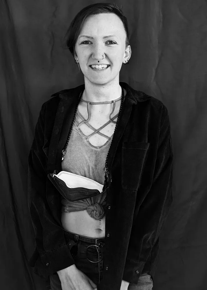 A person with shaved head, wearing a black jacket, smiling, with a nose ring and a stomach piercing.