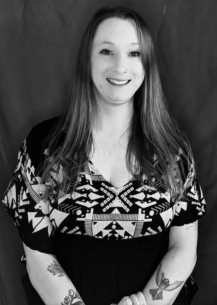 Woman smiling with long hair, patterned top and tattoos.