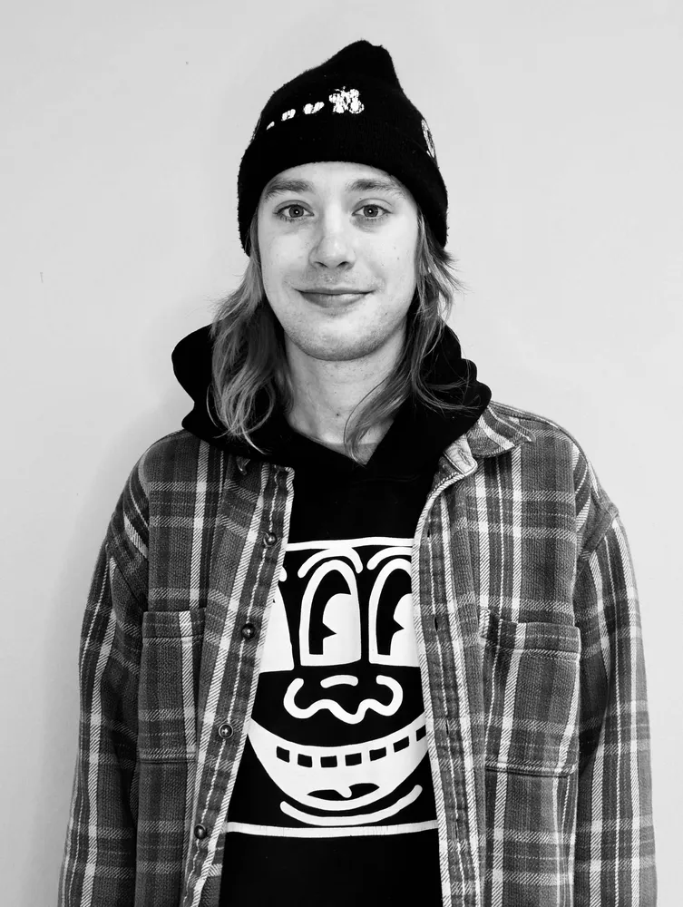 Young man wearing a beanie, hoodie with a graphic design, and plaid shirt, smiling.