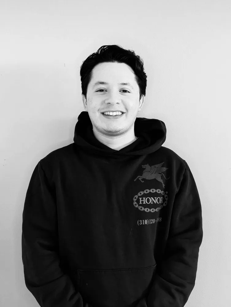 Smiling young man in a dark hoodie, standing in front of a plain wall.