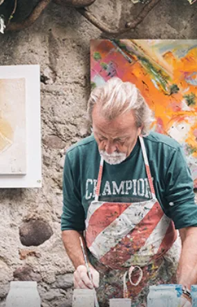 Artist in apron painting, surrounded by art in an old stone-walled studio.