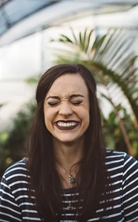 Woman with dark lipstick smiles broadly with eyes closed, striped shirt, and plant background.