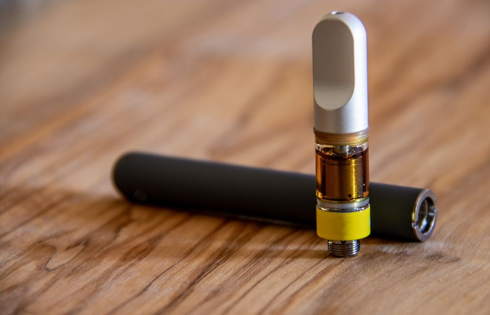 Vape pen with cartridge, dark gray body, sitting on wooden surface. Cartridge contains amber oil.