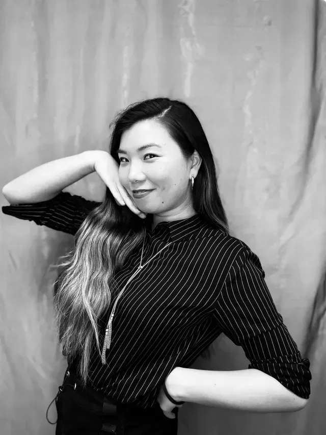 Woman with long hair poses, hand near face, in front of curtain.