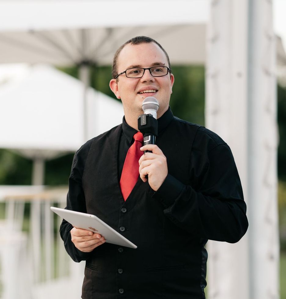 A man in a black shirt and red tie is holding a microphone and a tablet