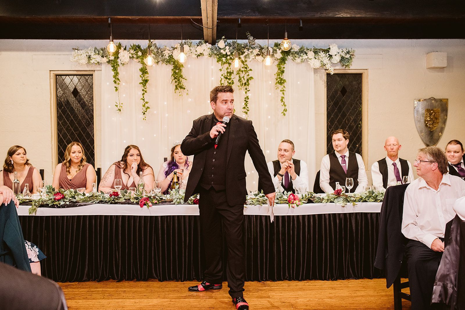 A man in a suit is standing in front of a microphone at a wedding reception.