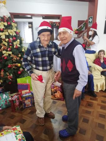 Dos personas con gorros de Papá Noel posando junto a un árbol de Navidad y regalos envueltos en el interior de una casa.