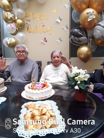 Dos personas sentadas en una mesa de cumpleaños con pastel, flores y globos dorados y negros sobre un fondo festivo.
