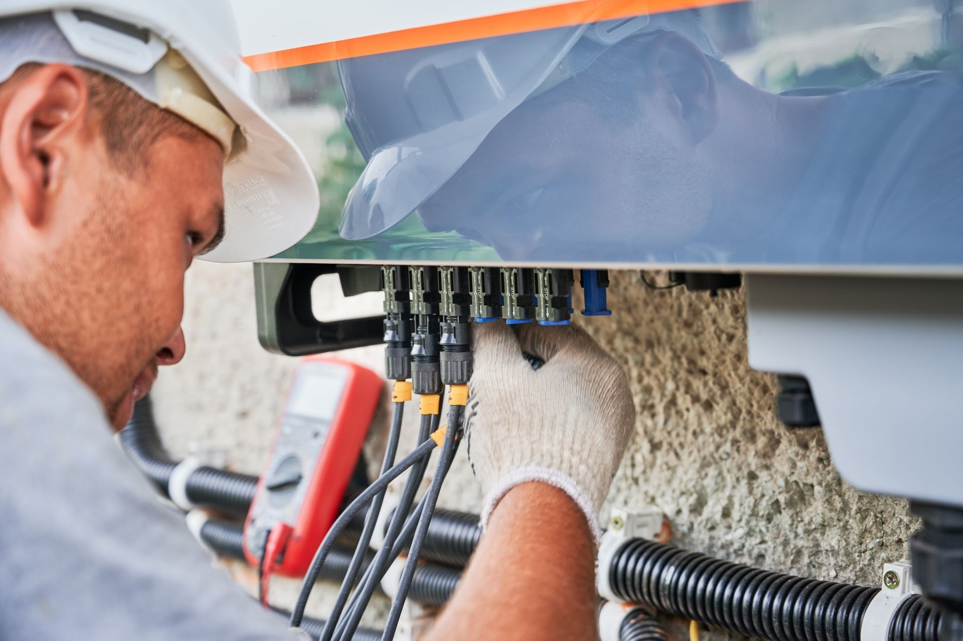 Solar panel technician connecting wires to an inverter on a wall, wearing gloves and a hard hat.