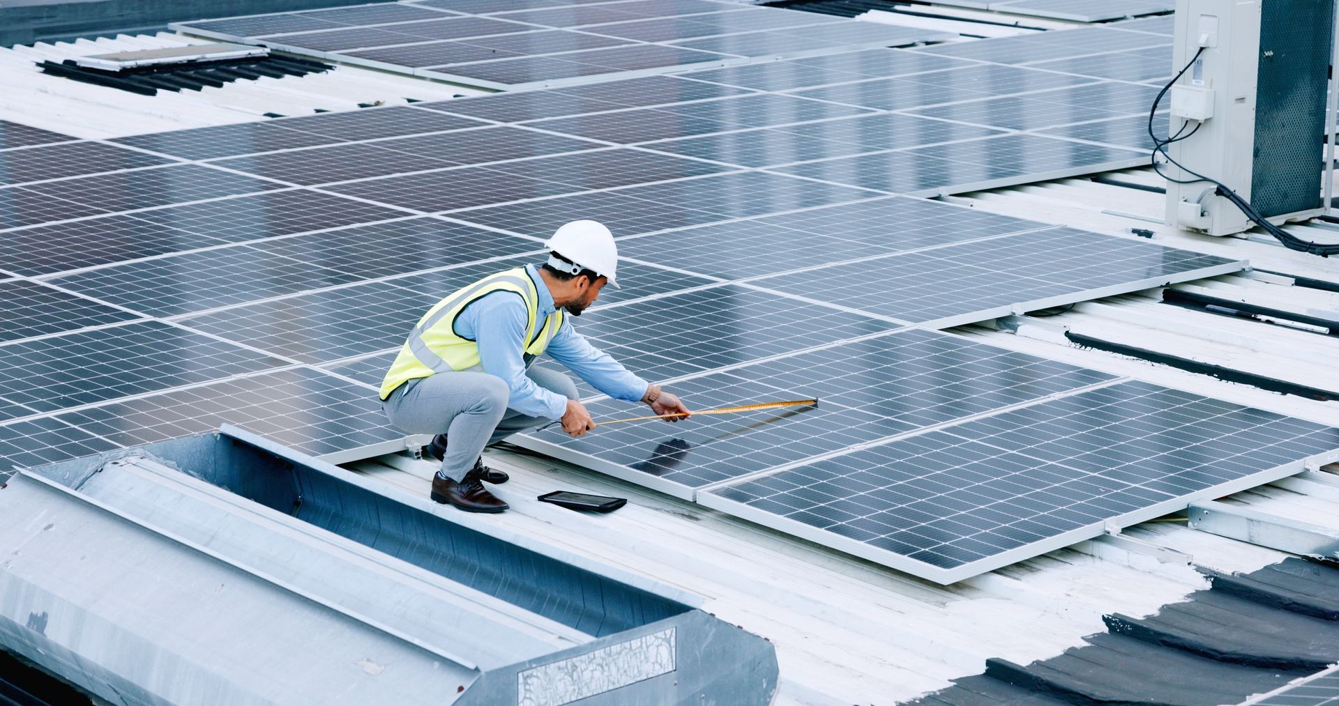 Solar panel technician inspecting panels on a rooftop, wearing a hard hat and safety vest.