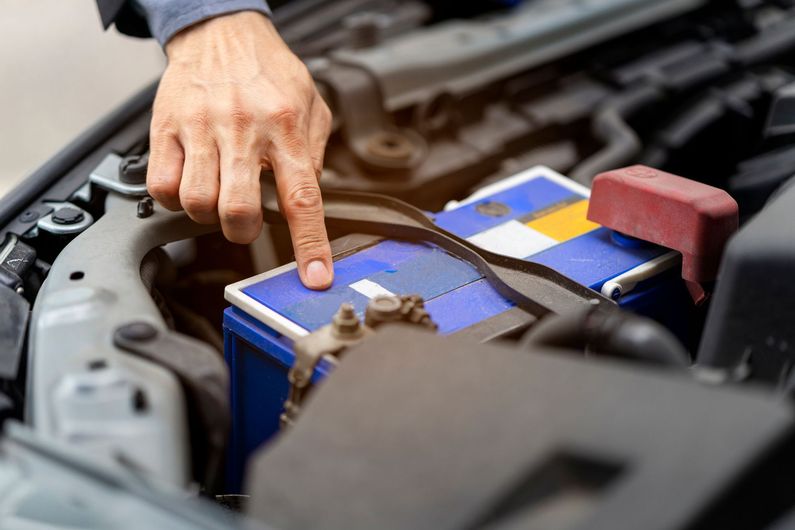 A person is pointing at a car battery under the hood.