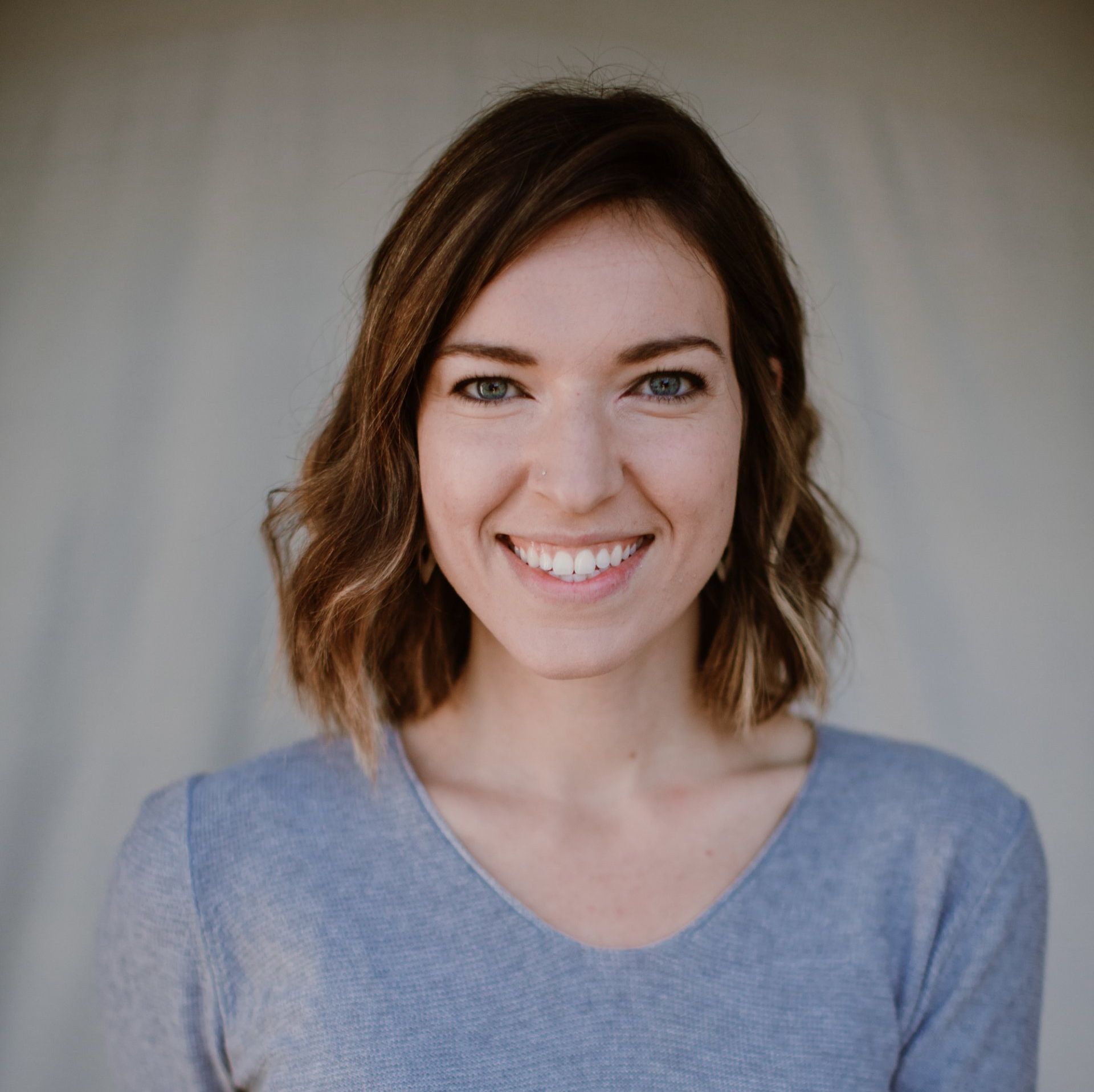 a woman with shoulder length brown hair with a gentle curl and highlights in a blue shirt smiles for the camera