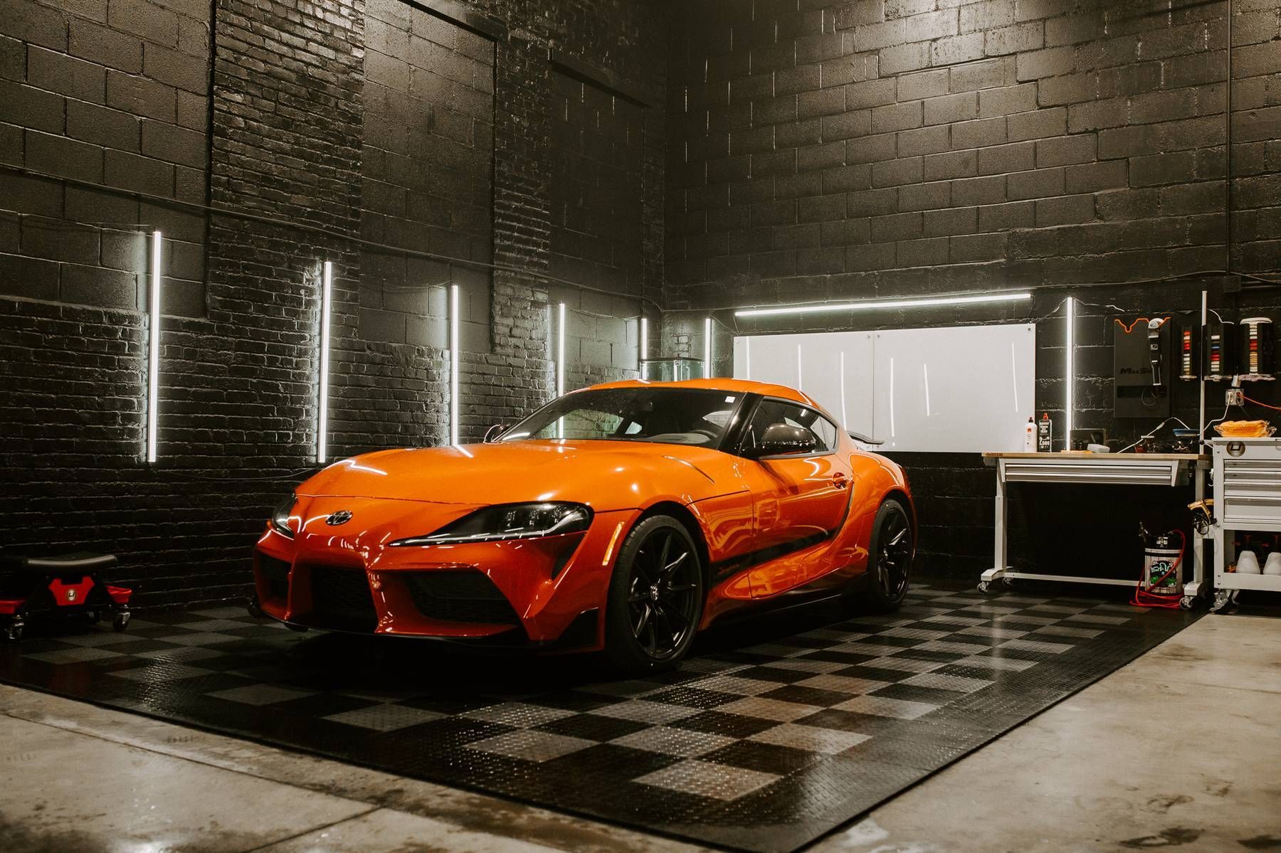 Orange sports car in a garage with black flooring and brick walls.