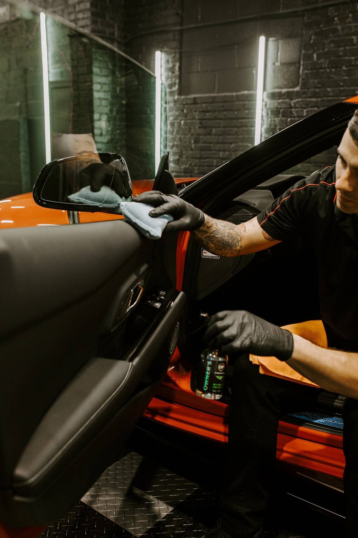 Man polishing an orange sports car in a garage, wearing gloves, using a power tool.