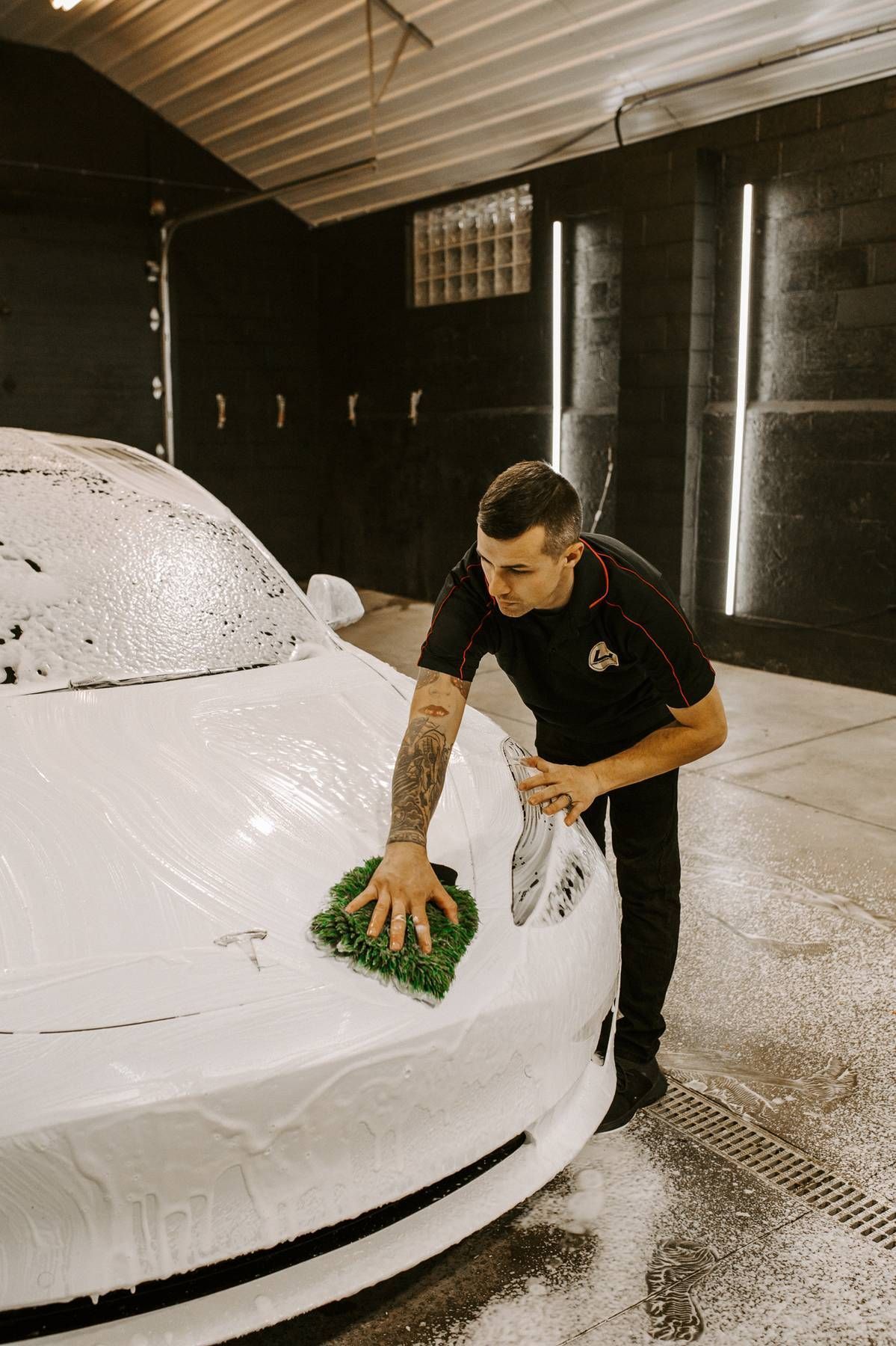 Man washes a white car with soapy foam, using a green sponge in a car wash bay.