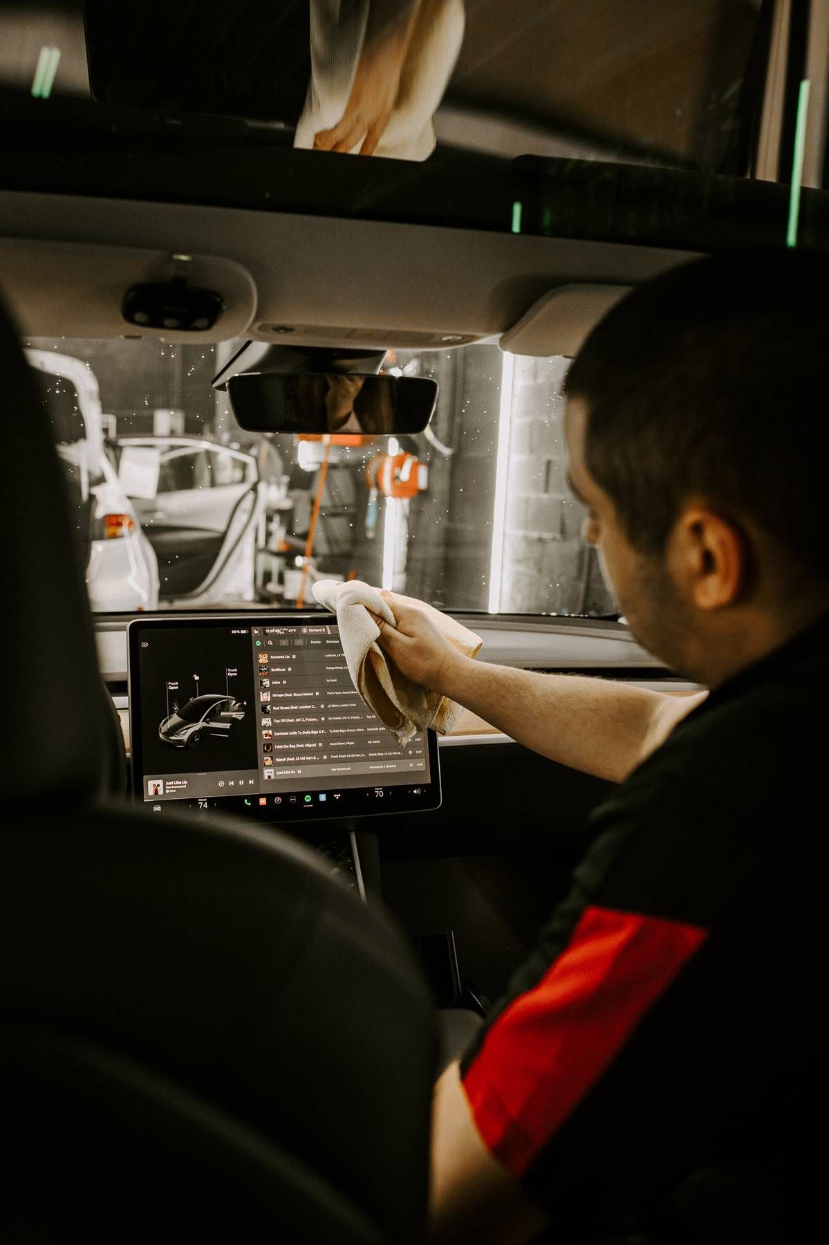 Man cleaning a car's touch screen inside the vehicle. He's wearing a black shirt with red accents. The interior is modern.