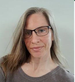 Jen is white and in her fifties. She has long brown hair and glasses and wears a gold / brown top.