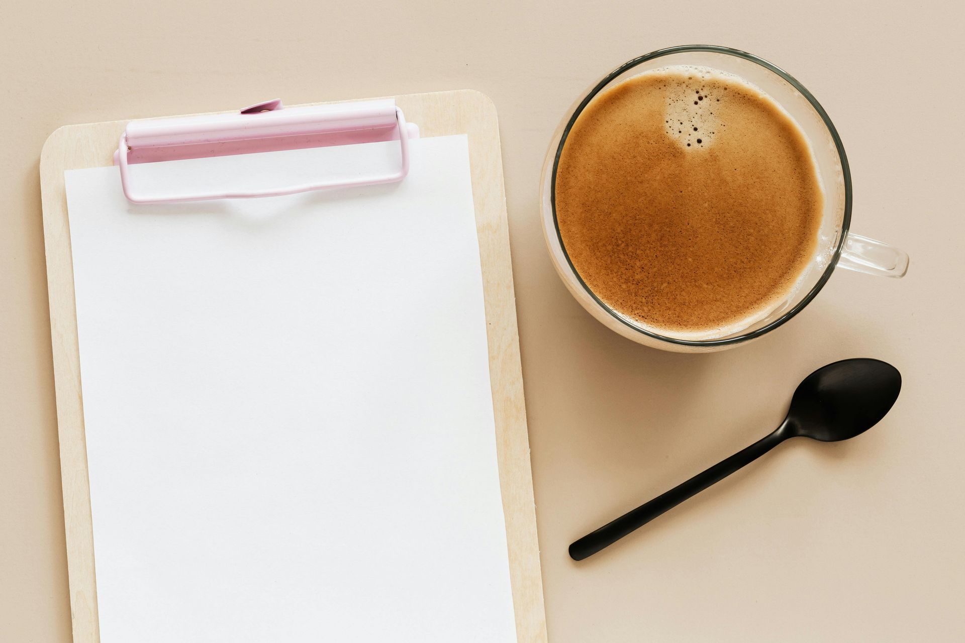 A clipboard with a blank sheet of paper, a frothy coffee and a black plastic spoon, viewed from above. 