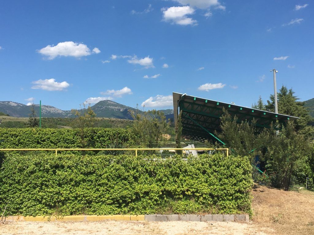 Fogliame verde con una ringhiera gialla di fronte a una struttura con un tetto verde inclinato sotto un cielo azzurro, montagne sullo sfondo.
