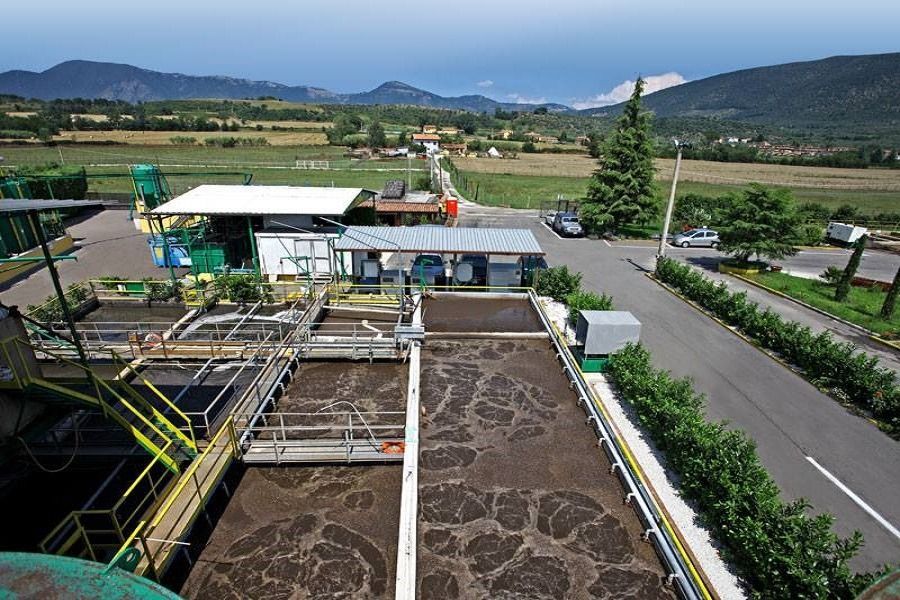 Impianto di trattamento delle acque reflue in un ambiente rurale con cisterne, edifici e una strada, con montagne sullo sfondo.