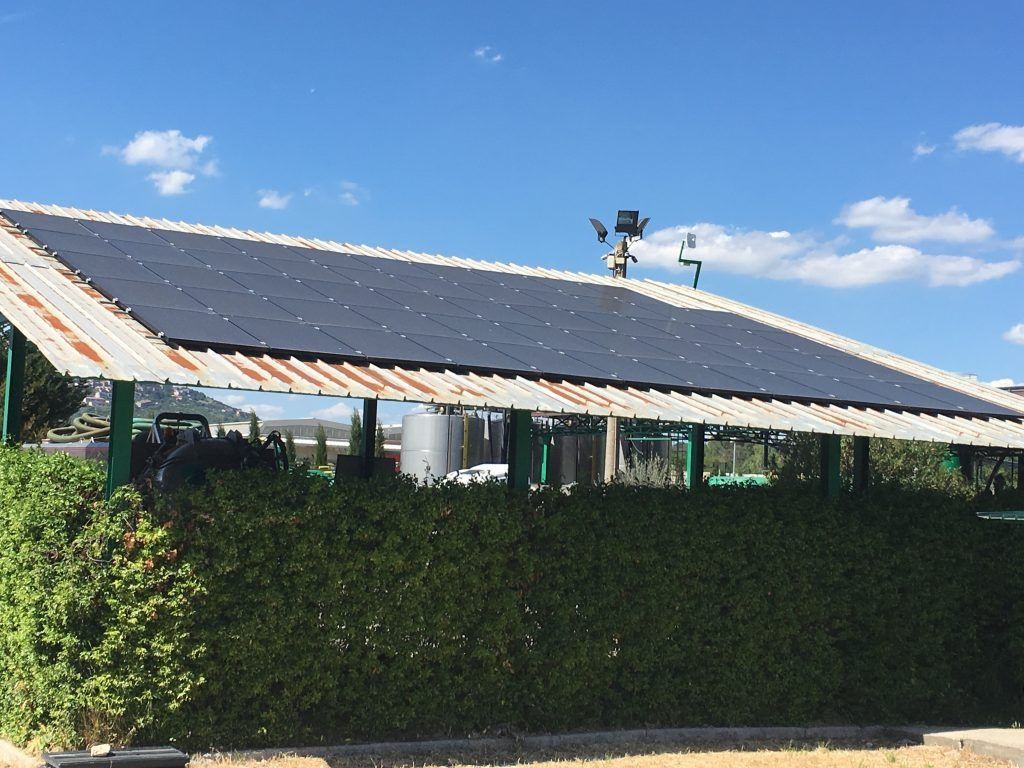Pannelli solari su un tetto di lamiera ondulata, che ricoprono una struttura con una siepe verde di fronte, sotto un cielo azzurro.