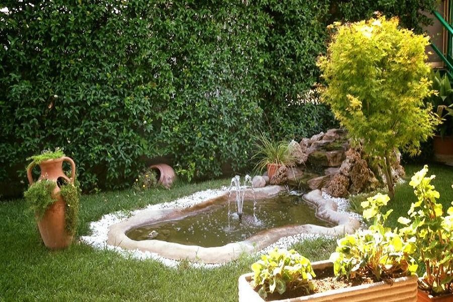 Un piccolo giardino con uno stagno, una fontana, del verde e un vaso ornamentale.