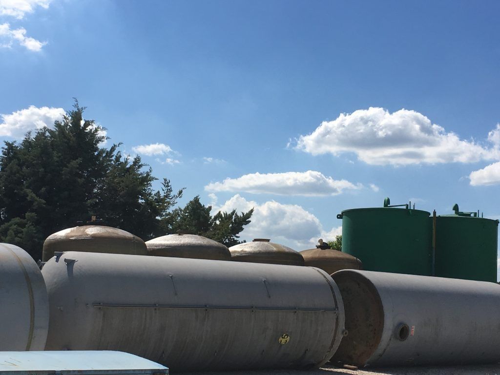 Serbatoi e container in una giornata di sole. Serbatoi grigio chiaro in primo piano, serbatoi verdi sullo sfondo, cielo azzurro con nuvole.