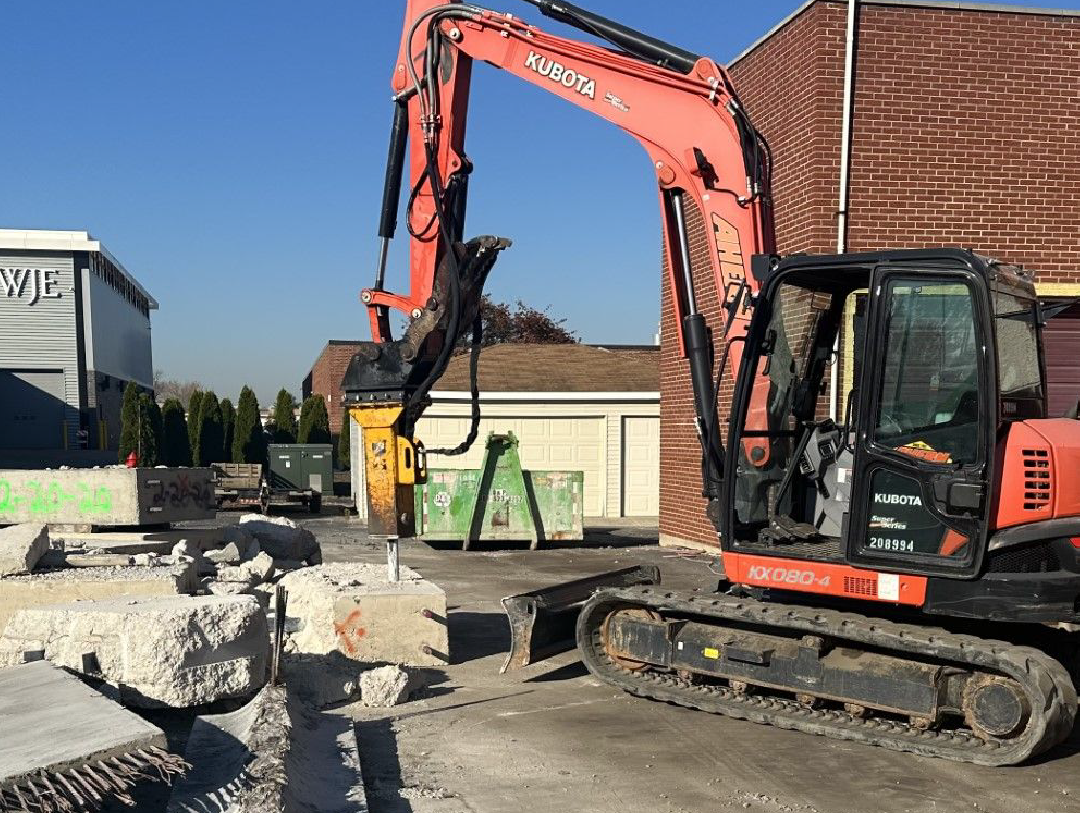 A red and black excavator is sitting in front of a brick building.