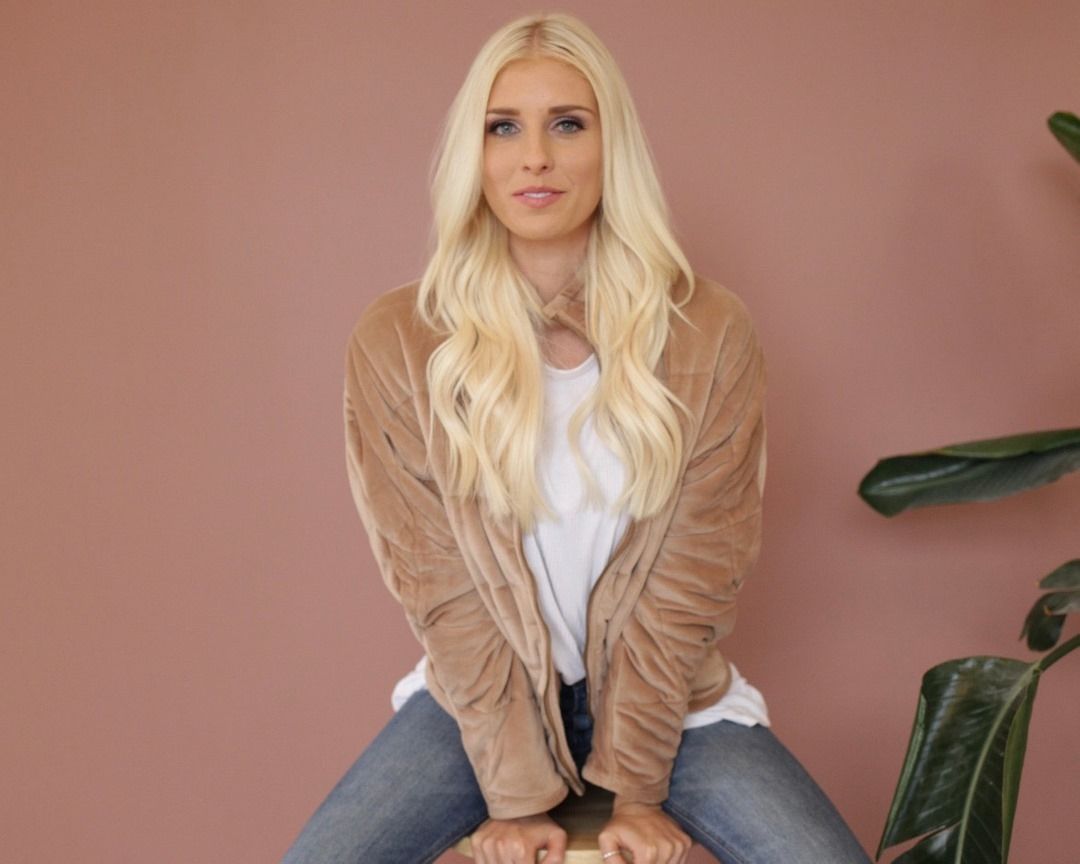 A blonde woman is sitting on a stool in front of a pink wall.