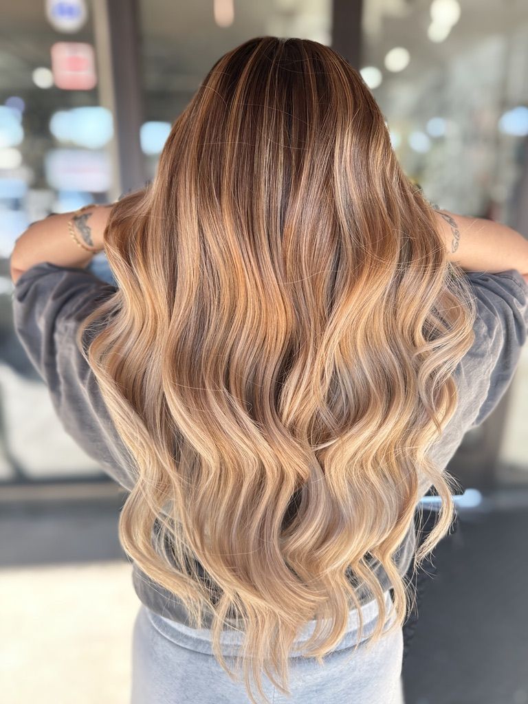 A woman with long blonde hair is standing in front of a building with her hands behind her head.