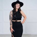A woman in a black dress and hat is standing in front of a white wall.