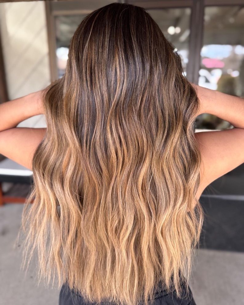 A woman with long brown hair is standing in front of a window with her hands behind her head.