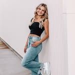 A woman in a black crop top and blue jeans is leaning against a wall.