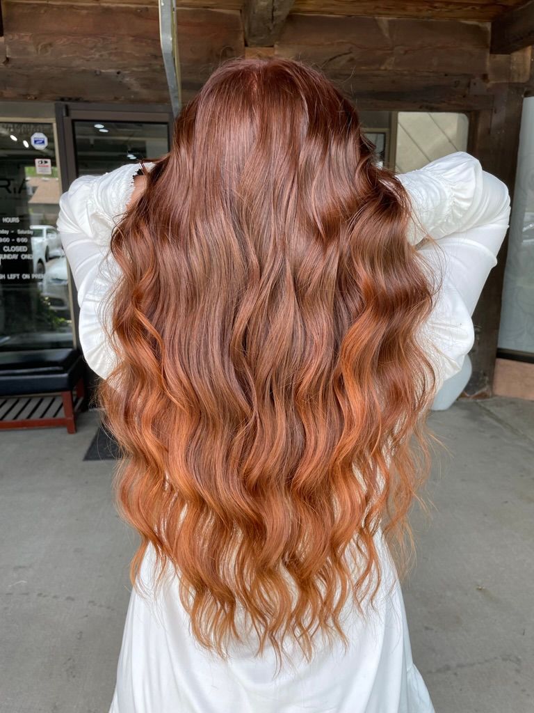 A woman with long red hair is standing in front of a building with her hands in her hair.