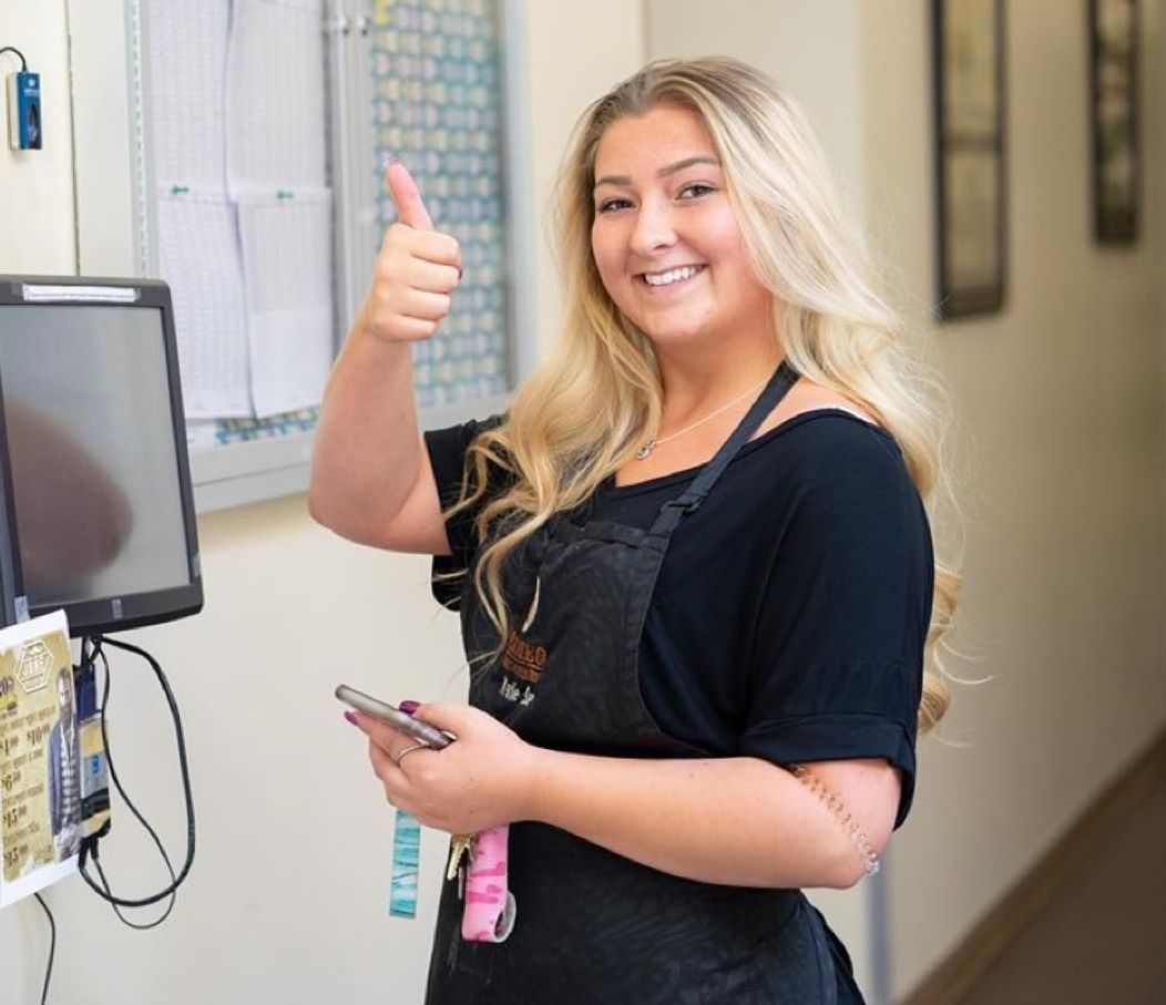A woman in an apron is giving a thumbs up while holding a cell phone.