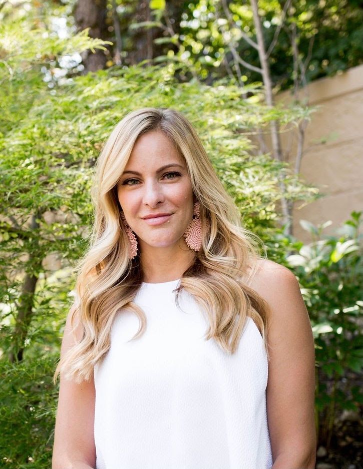 A blonde woman is sitting on a stool in front of a pink wall.