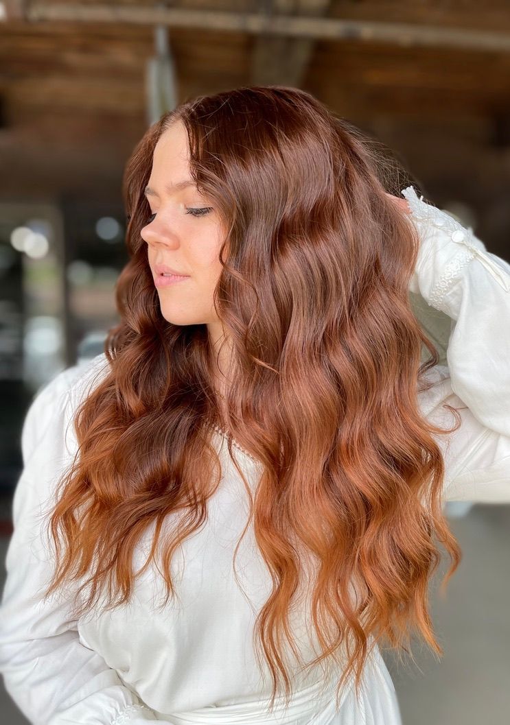 A woman with long red hair is wearing a white dress and holding her hair.