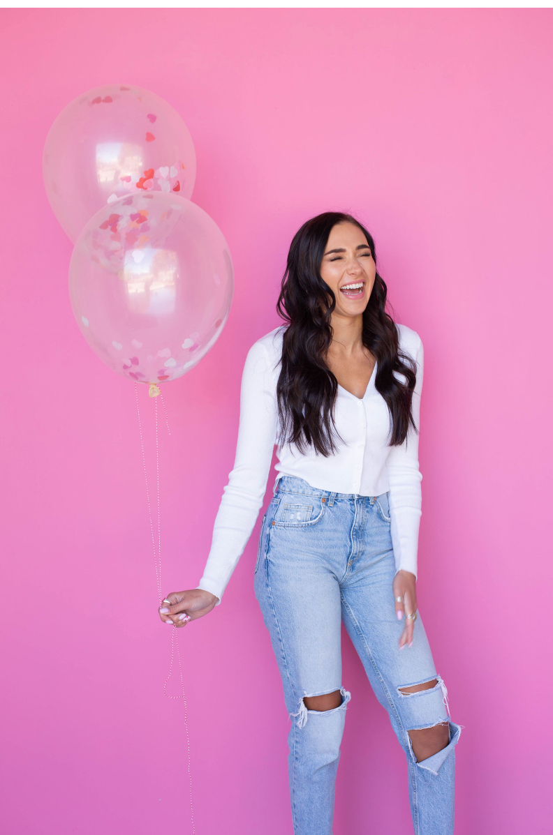 A woman is holding two balloons in front of a pink background.
