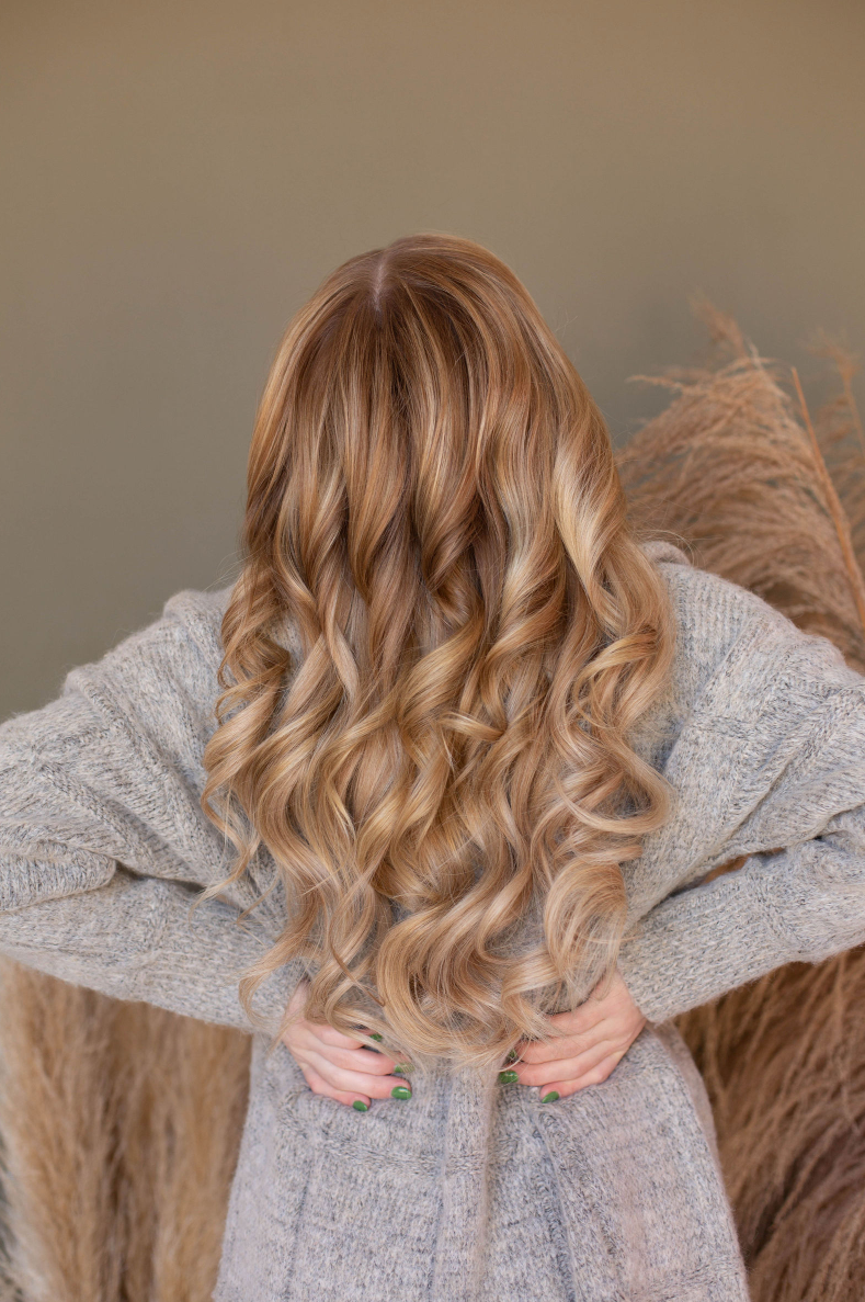 A woman with long blonde curly hair is wearing a grey sweater.