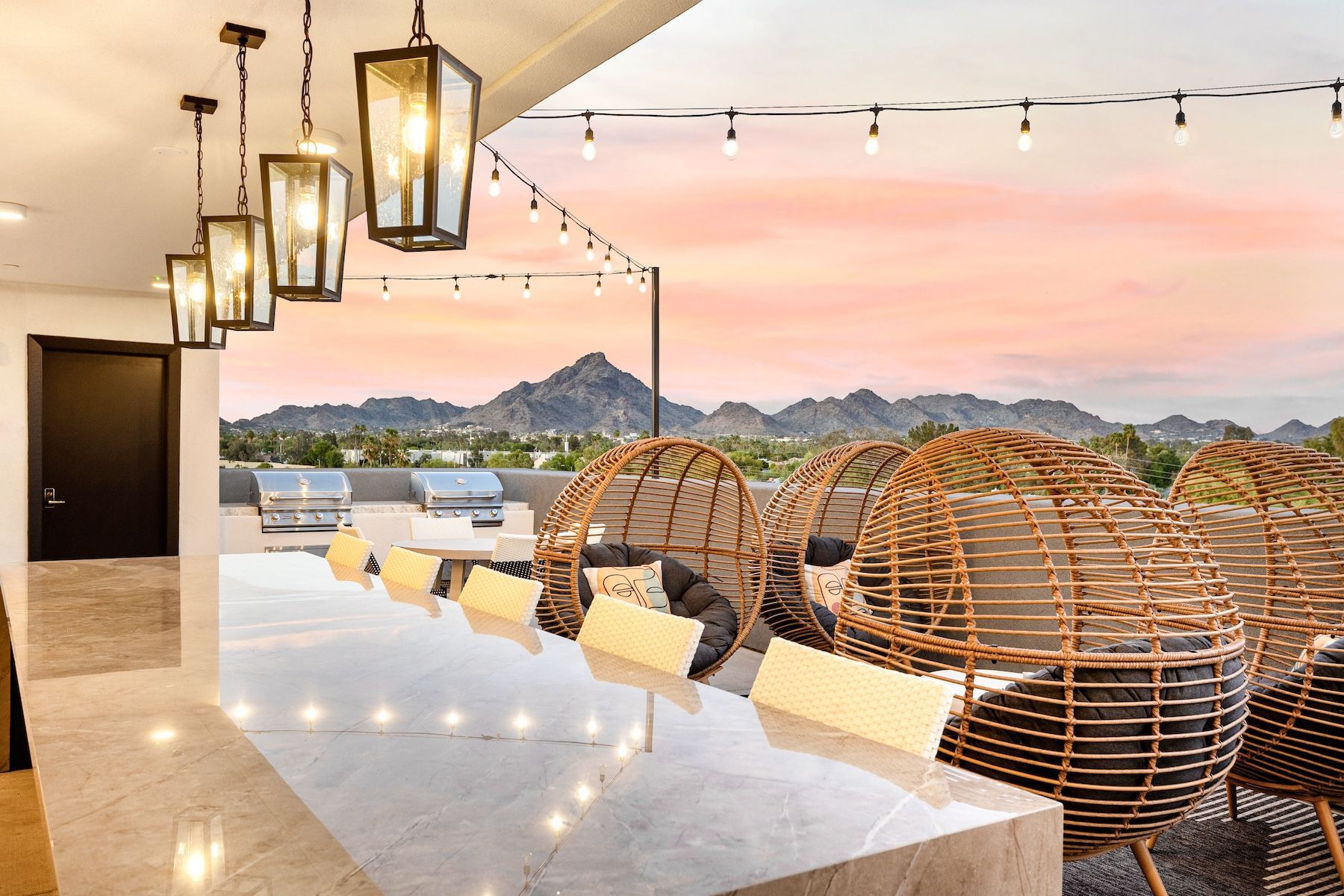 Rooftop patio with city views and hanging lights. Features bar, chairs, and wicker lounge chairs at sunset at Echo Biltmore in Phoenix, AZ..