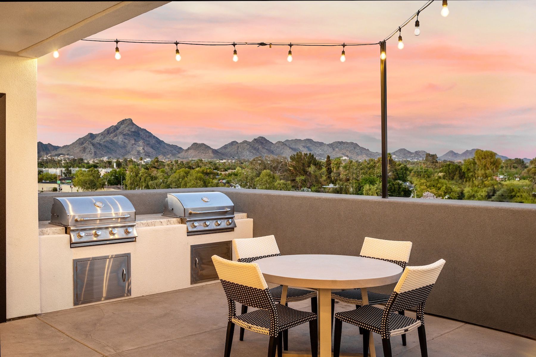 Rooftop patio with grills, a table, and chairs, overlooking mountains at sunset. String lights overhead at Echo Biltmore in Phoenix, AZ..