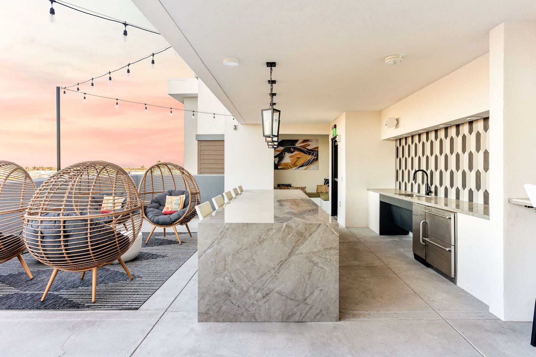 Rooftop patio with stone bar, seating, outdoor kitchen, string lights against a sunset sky at Echo Biltmore in Phoenix, AZ..
