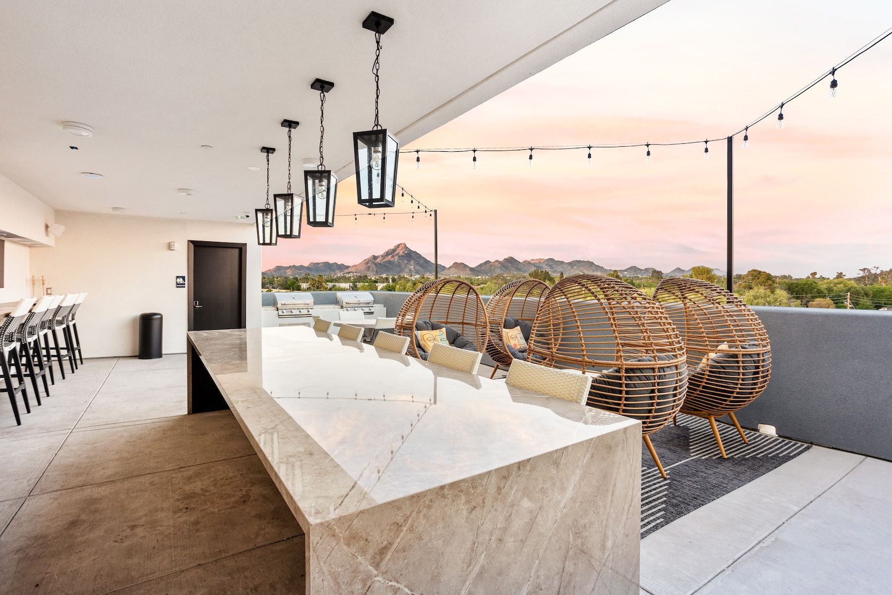 Rooftop patio with a long marble table, hanging lights, chairs, and a mountain view at sunset at Echo Biltmore in Phoenix, AZ..
