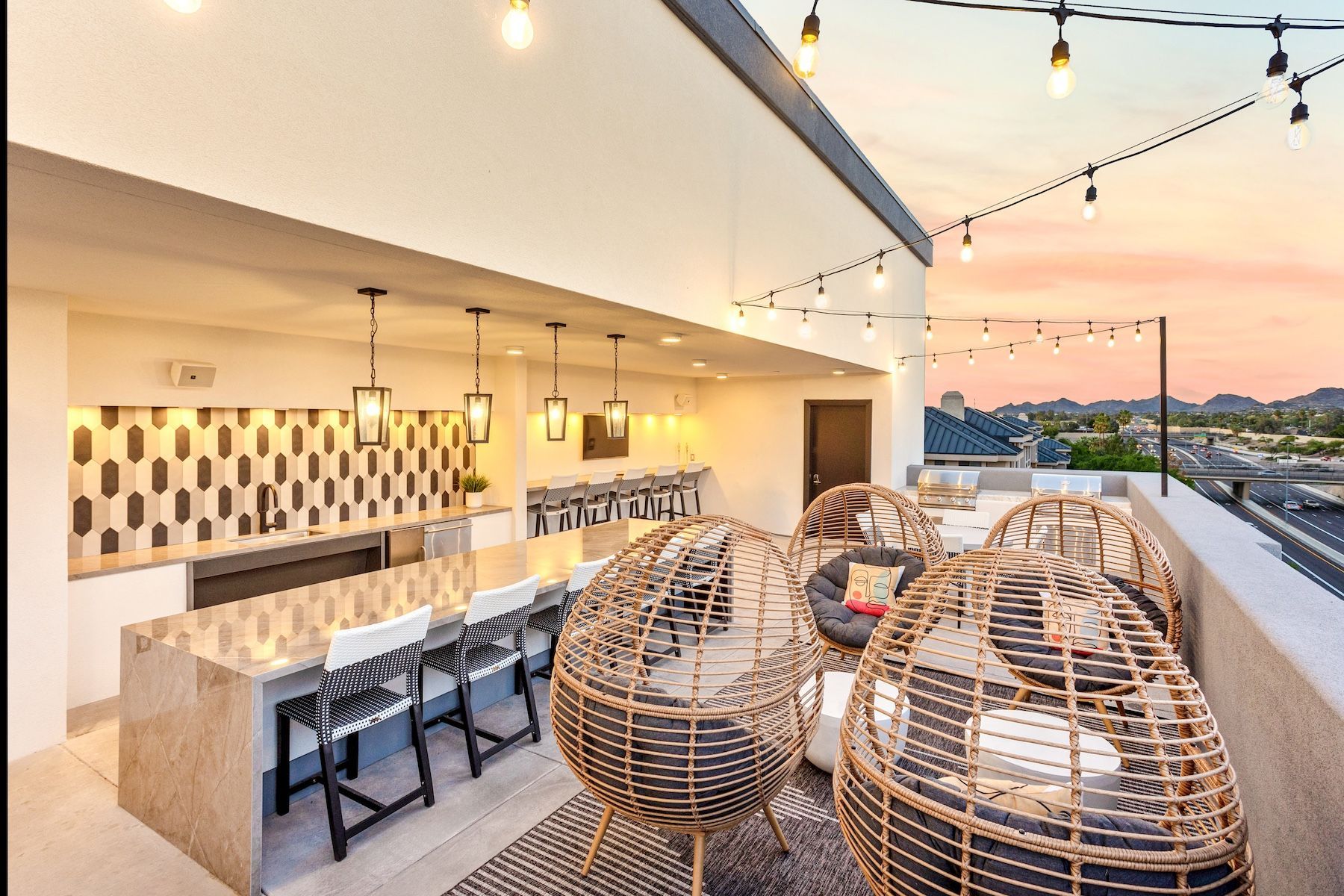Rooftop lounge with egg-shaped chairs, bar, string lights, and a city view at sunset.